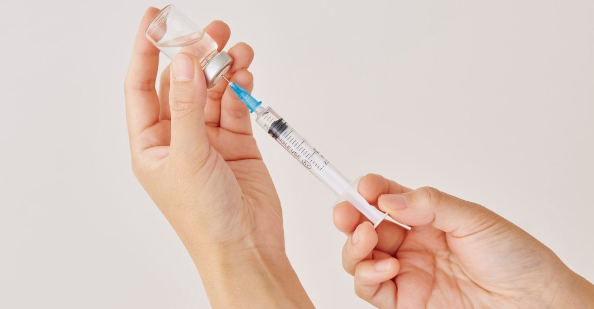 Mani che prelevano un farmaco da una fiala in vetro utilizzando una siringa graduata, su uno sfondo bianco e pulito