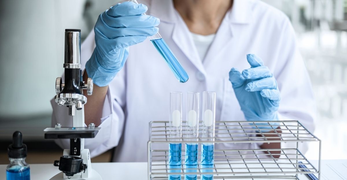 Ricercatrice in camice bianco e guanti azzurri che maneggia una provetta con liquido blu accanto a un microscopio da laboratorio