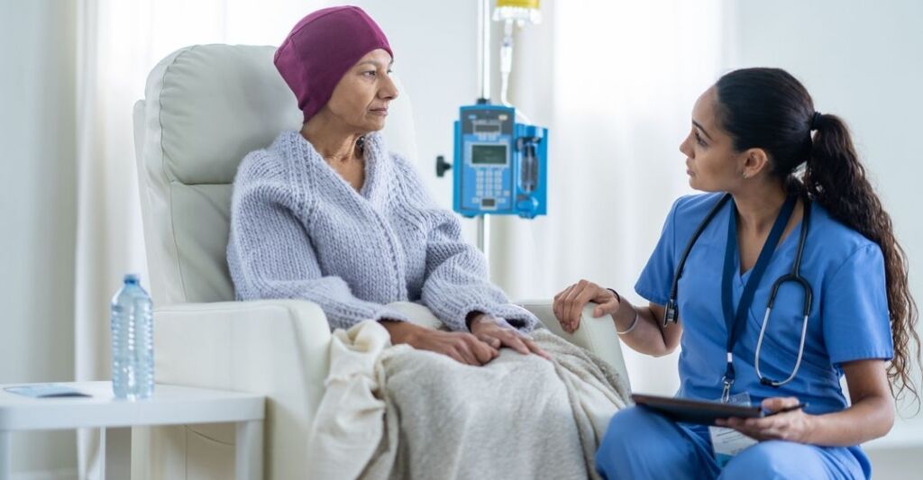 Una paziente con copricapo bordeaux seduta su una poltrona medica mentre parla con un'infermiera in divisa blu, con un supporto per infusione accanto
