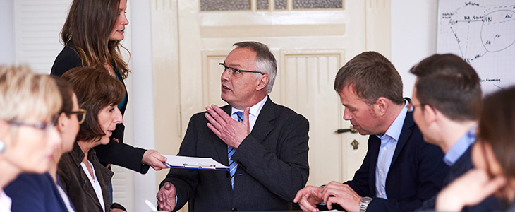 730x300 - Gruppe sitzt am Tisch, Frau reicht ein Klemmbrett an