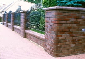 Ciepłobrązowe ogrodzenie ładnie zamyka zieloną ścianę ogrodu. Mur z klinkierowych cegieł połączony z elementami kutymi to dobry wybór dla tradycyjnej architektury. Na zdjęciu: cegła Adelajda, wymiary 240 x 75 x 40 mm, cena od 2,26 zł/szt. RÖBEN
