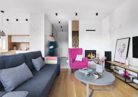 Wśród szarości, beżów i bieli wybija się amarantowy fotel Pola z Moma Studio. Z tego samego sklepu są kanapa Cuba i stolik kawowy Torring 90. W tle minimalistyczny kominek z pojemną wnęką na opał (wkład z kratki.pl).