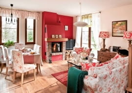 <B>Zgoda: salon, w którym przyjmujemy gości, musi wyglądać reprezentacyjnie. Ale to wcale nie znaczy, że ma być sztywny i oficjalny! Proszę tylko popatrzeć na prezentowane przez nas wnętrza. One dobitnie udowadniają, że elegancja może iść w parze z miłym, domowym klimatem.</B> <BR />MIEJSCE NA RELAKS. Łącząc naturalne materiały z ciepłymi barwami i meblami o ponadczasowych formach, stworzono apetyczny wystrój, który nie znudzi się zbyt szybko. Nutki słodyczy dodały mu tkaniny w kwiatowe wzory. Gdy kiedyś zostaną wymienione, smak tej mieszanki może być już całkiem inny. <BR />Kąt z kominkiem to bardzo ważne miejsce w tym salonie. Jego rangę podkreśla ściana pomalowana na złamany odcień czerwieni, do której konsekwentnie dobrano odcienie wzorów na poduszkach, abażurach lamp oraz pokrowcach przykrywających krzesła i kanapę. Mocne działanie tej barwy tonują duże płaszczyzny kremowej bieli (ściany) oraz jasnego brązu (drewniana podłoga i meble).