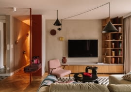 Salon wyposażono w welurową, jasnozieloną sofę z Etap Sofa, różowy fotel Chierowskiego marki 366 Concept oraz czarne stoliki kawowe Tamo. Podłogę zdobi dywan ze sklepu Rugvista. Strefę doświetlają wiszące nad kompozycją mebli czarne lampy od Kolorowe Kable i dwa kinkiety od ENOstudio. Ścianę z telewizorem wykończono strukturalnym tynkiem w beżowym odcieniu.