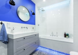 BIEL PŁYTEK I SZAROŚĆ MEBLI ożywia farba w odcieniu indygo, którą pomalowano fragment jednej ze ścian. Łazienkę oświetlają listwy LED zainstalowane pod wanną i nad nią (za załomem podwieszanego sufitu), halogeny zamontowane w suficie oraz kinkiety (Kartell) przy lustrze./fot. mat. red.