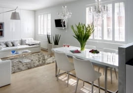 Wyspa - główne miejsce pracy - została przesłonięta nieco od niej wyższym murkiem. Ponieważ nie zasłonił on całkowicie mebli kuchennych i wysokiej zabudowy, wybrano szafki z frontami w kolorze ściany, co wtopiło je w tło. Aby nic nie rozbijało ich gładkiej płaszczyzny, zdecydowano się na szafki otwierane dotykowo. Harmonię we wnętrzu podkreślają użyte materiały wykończeniowe - na podłodze, tak jak w pozostałej części pomieszczenia, leżą panele, a ścianę we wnęce nad zlewem pokryto dekoracyjnym tynkiem strukturalnym. Przy murku od strony salonu znajduje się część jadalna. Blat sporego stołu jest zrobiony z takiego samego materiału co fronty szafek - płyty lakierowanej na wysoki połysk.