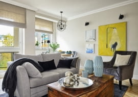 OKNA w pokoju dziennym wychodzą na południowy wschód, dlatego oprócz ozdobnych rolet rzymskich z lnu (Lniany Zaułek) są też rolety nieprzepuszczające światła. Obrazy gospodyni wybrała w galerii internetowej alesztuka.com. Autorem dużego obrazu z psem jest Filip Pręgowski, dwóch mniejszych Agata Kosmala. Poduszki i dywan są ze sklepu twojerolety.pl, kanapa z Furninova, a fotel - z TK Maxx. Niebieskie wazony i tacę z lustrzanymi kulami kupiono w Decoretro.