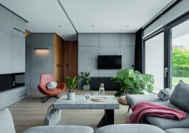 Wnętrza prezentowanego domu podążają za architekturą zewnętrzną z jej nowoczesną, jasną bryłą podkreśloną grafitowym dachem, garażem i oknami. W środku zastosowano podobne, jasno-ciemne zestawienie kolorów: niemal czarne dodatki (RAL 7021) z szarością i bielą. Do chłodnej kolorystyki dodano ciepłe drewno.