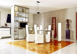 W otwartej części dziennej umowny zasięg jadalni wyznacza prostokąt z drewna merbau wkomponowany w posadzkę z żywicy epoksydowej. W tle - odsłonięta ściana z żelbetu. Nowoczesny regał ze stali nierdzewnej i matowego szkła sięga do samego sufitu, ale dzięki przeszkleniom wygląda lekko. Na półkach, otwieranych od strony kuchni, są przechowywane ozdobne naczynia; od strony jadalni zainstalowano telewizor.