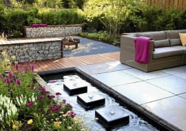 Small garden with wicker sofas on decked and paved patio, backed by Fargesia murielae - Bamboo hedge. Rectangular pond with row of square water features