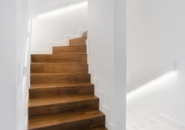 Betonowe schody zabiegowe prowadzące na piętro zostały obłożone drewnem. Zastosowanie takiego materiału w ciemnym kolorze w połączeniu z bielą ścian sprawia, że ten fragment wnętrza jest elegancki i nowoczesny zarazem