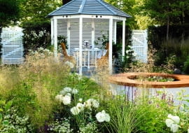 Table and chairs in wooden pavilion, outdoor dining, raised circular pond, Deschampsia cespitosa SLOWA KLUCZOWE: rose flower grasses perennial summerhouse gate pond seating furniture The Sadolin Refresh and Revive Garden RHS Hampton Court Flower Show 2010 Kathryn Hibberd June Summer small town romantic designer horizontal wide shot sunny white