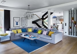 W części dziennej, zajmującej niemal cały parter, toczy się życie rodzinne i towarzyskie. Schody ze szklaną balustradą (Stol-Art) dzielą ją na dwie strefy: kuchenną oraz wypoczynkową. W części wypoczynkowej króluje szara narożna kanapa (Primavera Furniture), którą ożywiają poduszki z pakamera.pl.