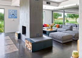 Wypoczynkowy obszar strefy dziennej wytycza dwustronny kominek - geometryczna forma z delikatnie ryflowanego betonu. Szarą paletę wnętrza dopełnia obraz Marii Widelak.