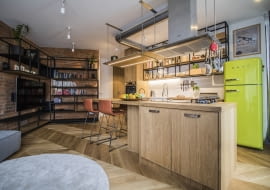 Otwarta kuchnia połączona jest z częścią wypoczynkową, którą umownie zaznaczono czerwoną cegłą. Na jej tle okazale prezentują się charakterystyczne dla stylu industrialnego metalowe meble.