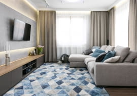 Strefa dzienna wypełniona jest naturalnym światłem dzięki dużym oknom. Wrażenie to potęgują podłogi z jasnego drewna i ściany pomalowane na jasno-szary kolor. Graficzne akcenty i niebieskie poduchy ożywiają monochromatyczną stylizację.