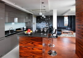 Widok katedry w Sewilli przesłoniętej rozłożystym drzewem uprzyjemnia gospodarzom wypoczynek przez cały rok. Kolorystyka fototapety idealnie pasuje do odcieni zasłon i mebli, jednocześnie dodaje wnętrzu przestrzeni. <BR /> MIESZKANIE. Wystrój części dziennej jest bardzo spójny. Barwa kuchennych frontów z lakierowanej na wysoki połysk płyty MDF nawiązuje do odcienia tkanin w salonie. Obudowę wyspy i fronty wysokiej zabudowy (widocznej po prawej stronie) zrobiono z palisandru. Na podłodze - gres i parkiet przemysłowy z palisandru. Główny blat roboczy oświetlają halogeny zamontowane w częściowo podwieszonym suficie, wyspę - efektowne lampki wiszące, kąt wypoczynkowy - LED-y zamontowane w wycięciach sufitu.