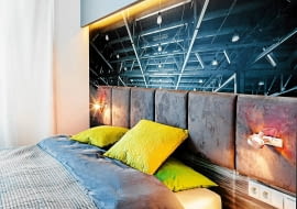 SYPIALNIA. Fototapeta, przedstawiająca surową stalową konstrukcję w opuszczonej hali fabrycznej, to po łóżku drugi element wystroju nawiązujący do klimatu loftowego. Kontrastem dla niej jest zagłówek z miękkich poduch obitych tkaniną przypominającą welur. Na podłodze ułożono dębowe deski, lekko pobielane. Pasuje do nich surowe drewno, z którego zrobiono łóżko. Gdy podstawa łóżka jest szersza od materaca, nie potrzeba już stolików nocnych. Niezbędne drobiazgi można postawić na brzegu mebla.