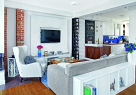 ZA KANAPĄ stanął niski regalik, w sam raz na czytane właśnie książki i dekoracyjne drobiazgi. Na granicy salonu i kuchni uwagę zwraca nietypowa półka na wino, zaprojektowana na wymiar i zrobiona przez stolarza.