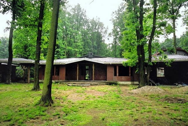 Sytuacja przed przebudową. Widok od frontu na nieproporcjonalnie niską bryłę domu otoczoną wysokimi drzewami