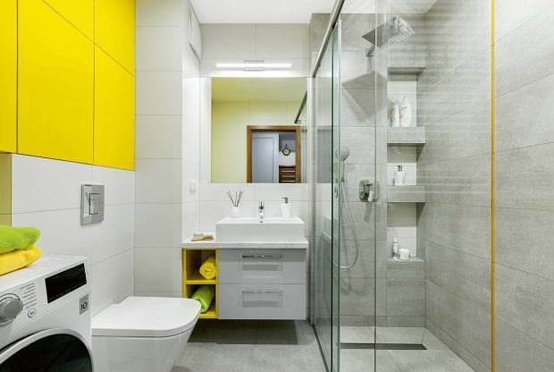 Wnękowe półki w kabinie prysznicowej celowo rozmieszczono na różnej wysokości - dzięki temu mogą z nich wygodnie korzystać i duzi, i mali. Warto dodać, że takie półki są bezpieczne i dla domowników (nie uderzą się o wystające elementy), i dla ustawionych na nich kosmetyków (nie spadną na podłogę). Na podłodze położono gresowe płytki Timbre Grey, na ścianach - Timbre White i Timbre cement; przy kabinie wkomponowano szklaną listwę Yellow (wszystko z firmy Tubądzin).