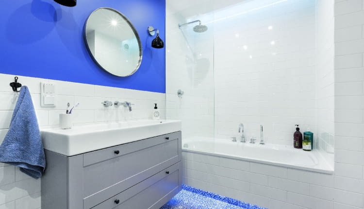 BIEL PŁYTEK I SZAROŚĆ MEBLI ożywia farba w odcieniu indygo, którą pomalowano fragment jednej ze ścian. Łazienkę oświetlają listwy LED zainstalowane pod wanną i nad nią (za załomem podwieszanego sufitu), halogeny zamontowane w suficie oraz kinkiety (Kartell) przy lustrze./fot. mat. red.