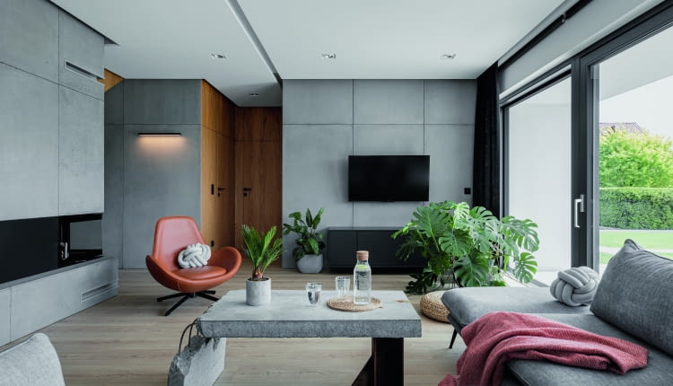Wnętrza prezentowanego domu podążają za architekturą zewnętrzną z jej nowoczesną, jasną bryłą podkreśloną grafitowym dachem, garażem i oknami. W środku zastosowano podobne, jasno-ciemne zestawienie kolorów: niemal czarne dodatki (RAL 7021) z szarością i bielą. Do chłodnej kolorystyki dodano ciepłe drewno.