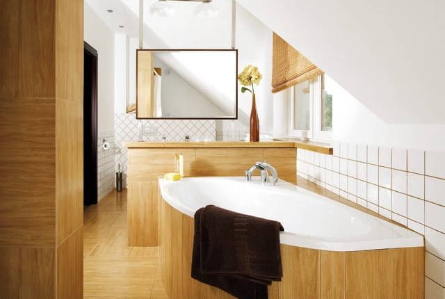 W UKRYCIU. Na pierwszy rzut oka ścianka wydzielająca bardziej intymną, kąpielową część pomieszczenia jest niemal niewidoczna. Nie dość, że idealnie wtapia się w tło, bo obłożono ją takimi samymi płytkami ceramicznymi jak podłogę, obudowę wanny i fragmenty ścian, to na dodatek od strony wejścia zasłaniają ją dwie szafki oraz zamontowana na jednej z nich spora umywalka. Chociaż płytki do złudzenia przypominają drewno, zapewniamy, że to jednak ceramika! Do wystroju wnętrza idealnie pasuje bambusowa roleta w oknie.