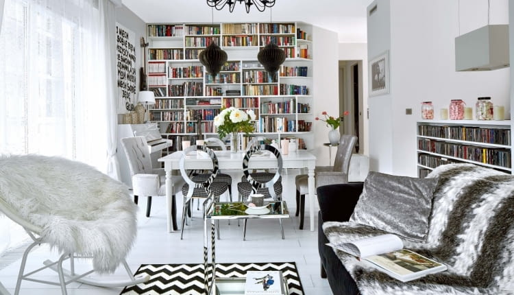 Wszechobecna biel: w tym kolorze są ściany, podłogi i większość mebli; właściciele przemalowali nawet pianino! Na takim neutralnym tle dobrze prezentuje się kolekcja książek, płyt i obrazów. Żyrandol, kupiony w jednym z marketów budowlanych, w innym wnętrzu mógłby wydać się pretensjonalny. Tu świetnie pasuje do stylizowanego stołu (IKEA) oraz srebrzystych krzeseł w stylu glamour. Głównym bohaterem salonu jest zrobiona na zamówienie biblioteczka. Boczne półki są ścięte, dzięki czemu tworzą spójną całość ze skośną ścianą.