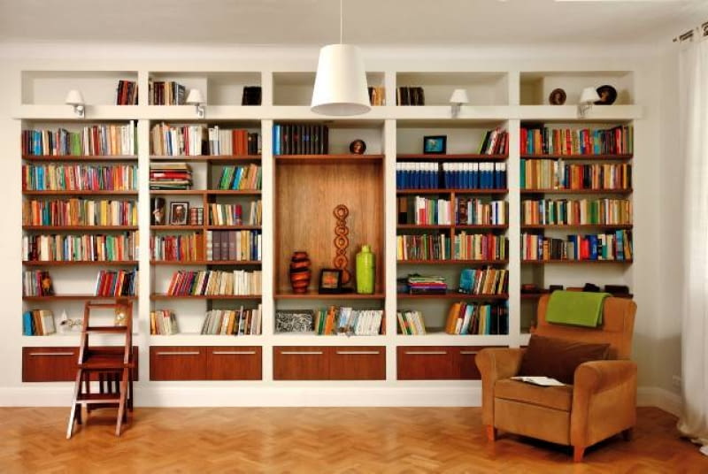 Aranżacje. Regał z płyt gipsowo-kartonowych zajmuje całą szerszą ścianę pokoju-biblioteki. Drewniane schodki umożliwiają sięganie po książki z wyższych półek. Wnętrze wydaje się jeszcze większe, niż jest w rzeczywistości. To dzięki jasnej kolorystyce z dominującą bielą w dwóch odcieniach (śnieżnym oraz kremowym), przełamaną barwą podłogi, półek i fotela. WNETRZA , POKOJ , BIBLIOTEKA