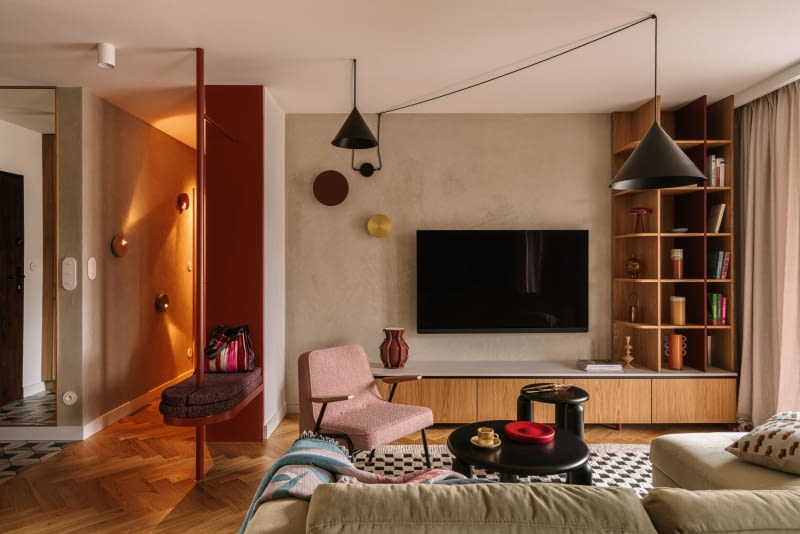 Salon wyposażono w welurową, jasnozieloną sofę z Etap Sofa, różowy fotel Chierowskiego marki 366 Concept oraz czarne stoliki kawowe Tamo. Podłogę zdobi dywan ze sklepu Rugvista. Strefę doświetlają wiszące nad kompozycją mebli czarne lampy od Kolorowe Kable i dwa kinkiety od ENOstudio. Ścianę z telewizorem wykończono strukturalnym tynkiem w beżowym odcieniu.