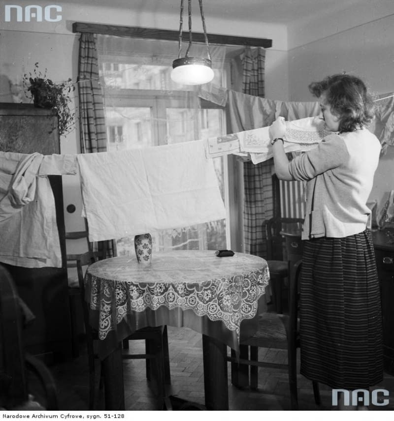 Polskie mieszkania w ciągu ostatnich 100 lat przeszły diametralne przemiany. W dwudziestoleciu międzywojennym wnętrza urządzane były z dużą starannością, meble i dodatki wybierano na lata, tak by służyły kilku pokoleniom. Nie było więc mowy o prowizorce. Ściany obficie zdobiono obrazami, grafikami, tkaninami, a czasem mapami i różnego rodzaju trofeami. Druga wojna światowa, a następnie niesprzyjający ustrój spowodowały, że naturalny rozwój stylu został na długo zahamowany. W mieszkankach Polaków zagościły dokładnie takie same meble i dekoracje. Meblościanki, tapczany, wersalki, boazerie, ale też polska ceramika były powtarzalnymi elementami w niemal każdym wnętrzu. Zmiana ustrojowa w 1989 roku otworzyła drzwi na nowe i przyniosła zachwyt zachodnimi wzorcami oraz nieposkromioną wolność w kreowaniu przestrzeni prywatnych. Dziś wciąż pozostajemy pod silnym wpływem zagranicznych stylów, jednak podchodzimy do nich bardziej krytycznie niż to miało miejsce jeszcze 10-15 lat temu. Obecnie typowe polskie mieszkanie to coraz częściej pokój połączony z kuchnią, współczesna wersja meblościanki z telewizorem w centralnej części i obowiązkowo jakiś mebel z IKEA. Jest to obraz zgoła odmienny od tego, co było w latach 20 i 30. </BR></BR> <B>Ok. 1955 r.: Kobieta rozwieszająca na sznurku w pokoju wypraną bieliznę.</B> polskie wnętrza, jak mieszkają Polacy