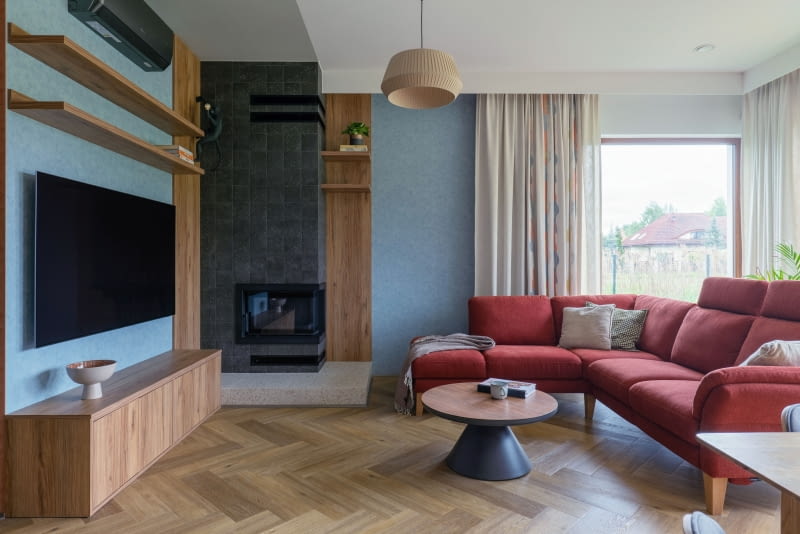 . Obok kominka stanęła duża, rodzinna sofa w bordowym odcieniu oraz stolik łączący drewniane i czarne elementy – zestaw spina całą strefę wypoczynkową.