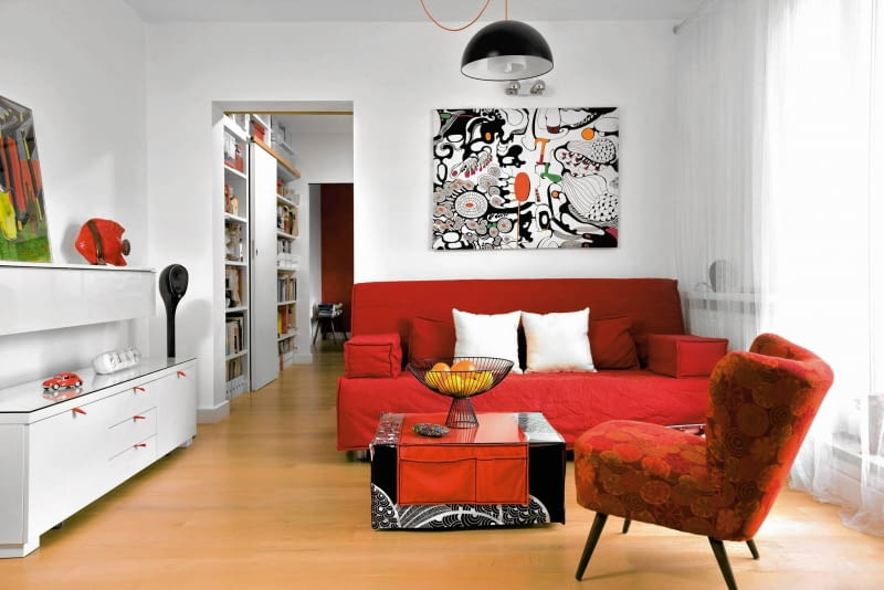 Z białymi ścianami kontrastują meble w mocnych kolorach - sofa (IKEA) i fotel z lat pięćdziesiątych w nowym obiciu (zdobycz z internetu). Abstrakcyjny obraz to po prostu kawałek czarno-białej tkaniny z IKEA, który gospodarze rozpięli na blejtramie i pokolorowali. Stolik kawowy jest dziełem projektantki; zrobiła go z płyty meblowej i 'ubrała' w pokrowiec i bieżnik z kieszeniami. Szufladom w białej komodzie dodano czerwone uchwyty z taśmy pasmanteryjnej - teraz łatwiej je otworzyć. aranżacja salonu, wystrój wnętrz