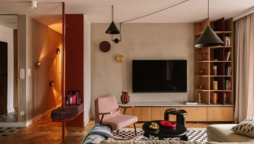 Salon wyposażono w welurową, jasnozieloną sofę z Etap Sofa, różowy fotel Chierowskiego marki 366 Concept oraz czarne stoliki kawowe Tamo. Podłogę zdobi dywan ze sklepu Rugvista. Strefę doświetlają wiszące nad kompozycją mebli czarne lampy od Kolorowe Kable i dwa kinkiety od ENOstudio. Ścianę z telewizorem wykończono strukturalnym tynkiem w beżowym odcieniu.