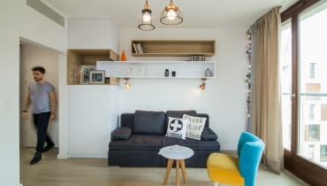 Kanapa rozkładana, musiała być dosyć prosta w formie, maksymalnie wygodna, no i musiała zmieścić się pomiędzy zabudową a oknem. W mebel słupek wkomponowano półki, które wprowadzają trochę rytmu i geometrii. Niewielki fotel i stolik utrzymane są w lekkiej stylistyce lat 50-60. Fotel w wyraźnej kolorystyce nadaje mieszkaniu barw i przewrotności. 30-metrowa kawalerka dla mężczyzny