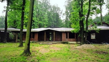 Sytuacja przed przebudową. Widok od frontu na nieproporcjonalnie niską bryłę domu otoczoną wysokimi drzewami