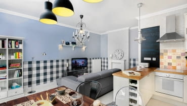 W pokoju dziennym nowoczesne elementy wystroju (narożna kanapa, lampa podłogowa) zgodnie egzystują z tradycyjnymi (żyrandol świecowy, drewniany regał, ręcznie malowany zegar, zdobiona drewniana obudowa kominka). Mieszanka stylowa panuje również w części jadalnej - przy rustykalnym stole stoją krzesła industrialne i pałacowe. I wszystko do siebie pasuje! Część wypoczynkową umownie wyodrębnia z pomieszczenia lamperia w starym stylu, którą zrobiono jednak nowoczesnym sposobem - dół ściany wyklejono tapetą i wykończono listwami sztukateryjnymi. aranżacje wnętrz, urządzanie wnętrz, mieszkanie