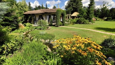 Strumyczek o naturalistycznie obsadzonych brzegach prowadzi w stronę krytej gontem altany otoczonej gęstwiną zieleni.