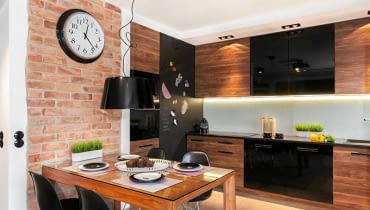 Zamiast jednego centralnego źródła światła - cztery podwójne halogeny (firmy Aquaform), zainstalowane w regularnych odstępach w podwieszonym suficie. Nie tylko rozjaśniają one całą kuchnię, ale także doświetlają blat roboczy. Większość szafek ma fronty fornirowane orzechem amerykańskim (z tego drewna jest również stół), pozostałe są z lakierowanej na czarno płyty MDF. Podłoga, wbrew pozorom, nie jest wyłożona drewnem, ale imitującym je gresem. Płytki o różnej szerokości ułożono 'na mijankę'. Przy cokołach szafek stojących zamontowano wodoodporną taśmę LED. Jej ciepłe światło jest efektowną ozdobą zwłaszcza po zmroku, gdy inne źródła światła w kuchni są wyłączone. Kuchnia, oświetlenie, meble kuchenne