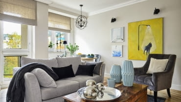 OKNA w pokoju dziennym wychodzą na południowy wschód, dlatego oprócz ozdobnych rolet rzymskich z lnu (Lniany Zaułek) są też rolety nieprzepuszczające światła. Obrazy gospodyni wybrała w galerii internetowej alesztuka.com. Autorem dużego obrazu z psem jest Filip Pręgowski, dwóch mniejszych Agata Kosmala. Poduszki i dywan są ze sklepu twojerolety.pl, kanapa z Furninova, a fotel - z TK Maxx. Niebieskie wazony i tacę z lustrzanymi kulami kupiono w Decoretro.