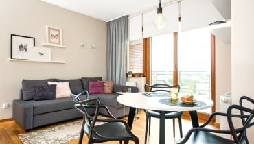 Jedną ze ścian pokoju niemal w całości zajmują przeszklone dwuskrzydłowe drzwi i okno balkonowe. Panoramę okolicy można więc oglądać, nie wstając z kanapy. Zasłona w rogu pełni jedynie funkcję dekoracji, dlatego zawieszono ja na krótkim karniszu. małe mieszkanie, salon, aranżacja salonu