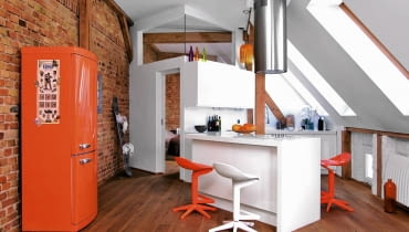Ulubiony kolor gospodarza? Nietrudno zgadnąć: pomarańczowy. Nie zabrakło go w aneksie kuchennym urządzonym przy sypialni. Taką barwę ma lodówka (firmy Ardo), stołki barowe i dodatki. Szafki zostały zrobione na wymiar z lakierowanej płyty MDF. Półwysep z płytą grzejną i piekarnikiem służy również jako miejsce do jedzenia. mieszkanie na strychu, skosy w mieszkaniu