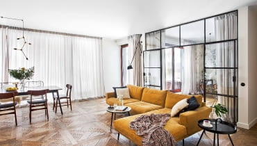 Strefę dzienną mieszkania wyodrębniono innym wzorem podłogi. Przy stole (BoConcept) komplet krzeseł vintage (Przetwory). Miodowa kanapa BoConcept, stolik kawowy Westwing.pl.