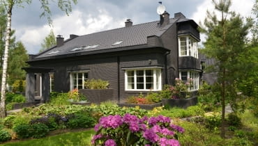 Widok na dom od strony ogrodu. Ciemna elewacja to niecodzienny sposób na wykończenie domu jednorodzinnego. W intensywnie zielonym otoczeniu sprawdza się jednak znakomicie, tworząc piękne tło dla starannie wypielęgnowanych roślin dom na wsi