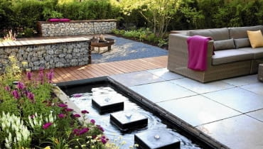 Projekty ogrodów. Gabiony wypełnione kamieniami dobrze izolują różne zakątki ogrodu Small garden with wicker sofas on decked and paved patio, backed by Fargesia murielae - Bamboo hedge. Rectangular pond with row of square water features