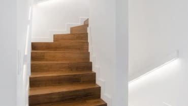 Betonowe schody zabiegowe prowadzące na piętro zostały obłożone drewnem. Zastosowanie takiego materiału w ciemnym kolorze w połączeniu z bielą ścian sprawia, że ten fragment wnętrza jest elegancki i nowoczesny zarazem