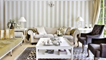 Ścianę pomalowano w szerokie szaro-beżowe pasy (podobny kolor mają obicia niektórych tapicerowanych mebli). Po obu stronach kanapy stoją stoliki pomocnicze; na nich wyeksponowano dekoracyjne przedmioty. Ścianę pomalowano w szerokie szaro-beżowe pasy (podobny kolor mają obicia niektórych tapicerowanych mebli). Po obu stronach kanapy stoją stoliki pomocnicze; na nich wyeksponowano dekoracyjne przedmioty.