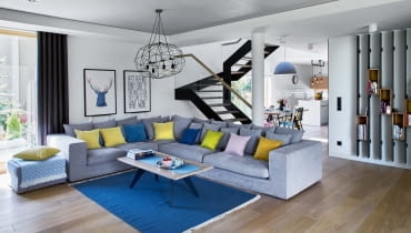 W części dziennej, zajmującej niemal cały parter, toczy się życie rodzinne i towarzyskie. Schody ze szklaną balustradą (Stol-Art) dzielą ją na dwie strefy: kuchenną oraz wypoczynkową. W części wypoczynkowej króluje szara narożna kanapa (Primavera Furniture), którą ożywiają poduszki z pakamera.pl.