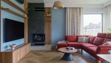 . Obok kominka stanęła duża, rodzinna sofa w bordowym odcieniu oraz stolik łączący drewniane i czarne elementy – zestaw spina całą strefę wypoczynkową.